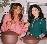 Two women stand in a kitchen behind brown cookware; one woman holds a large wooden spoon and both are smiling at the camera.