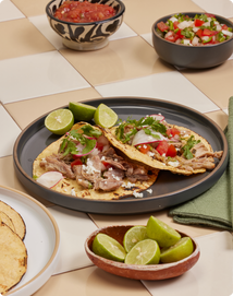 Two tacos with shredded meat, diced tomatoes, onions, and cilantro on a plate with lime wedges, surrounded by salsa, pico de gallo, and a bowl of lime slices.