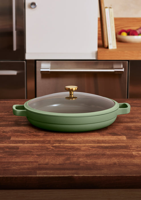 A green lidded skillet with a gold handle sits on a wooden kitchen counter, with books and a bowl of fruit in the background.
