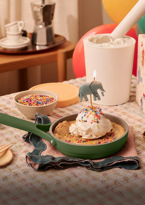 A skillet cookie topped with ice cream, sprinkles, and an elephant-shaped candle sits on a checkered cloth, with a container of marshmallows and bowls of sprinkles nearby.