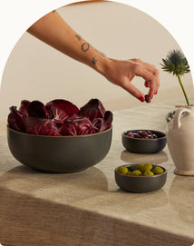 A hand reaches for a bowl of black olives on a table with a large bowl of radicchio, a small bowl of green olives, and a ceramic pitcher.