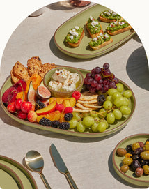 A platter with assorted fresh fruits, crackers, and cheese is on a table set with utensils. Other plates with appetizers and olives are also visible.