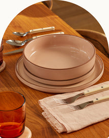 A set of pink ceramic dinnerware including a bowl and two plates is arranged on a wooden table with cutlery, a napkin, and an amber glass.