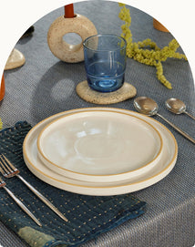 A neatly set table with two stacked plates, silverware on a blue textured napkin, a blue glass, two spoons, and decorative objects on a gray tablecloth.