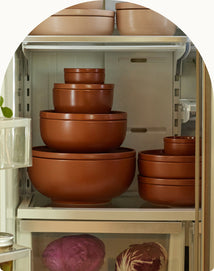 Several stacked, brown mixing bowls of various sizes are placed on a refrigerator shelf, with heads of cabbage visible in the lower compartment.