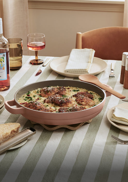 A pan of cooked chicken over creamy risotto sits on a striped tablecloth next to plates, napkins, a wine glass with rosé, and a slice of bread.