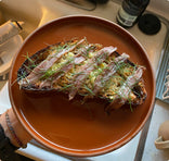 A hand holds a brown plate with a slice of toast topped with herbs and thin slices of cured meat, positioned over a kitchen sink.