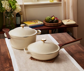 Two cream-colored cookware pieces with wooden handles, a pot with a lid and a pan with a lid, sit on a table runner in a kitchen setting with books and produce in the background.