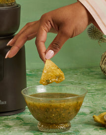 A hand dips a tortilla chip into a glass bowl of green salsa on a green marbled surface.