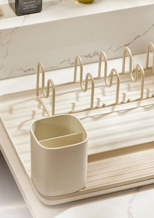 A beige dish rack with a divided utensil holder sits on a kitchen countertop with a marble backsplash and a bottle of olive oil in the background.