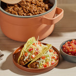 Two hard shell tacos filled with ground meat, shredded lettuce, and diced tomatoes are on a plate, with a bowl of salsa and a pot of cooked ground meat in the background.