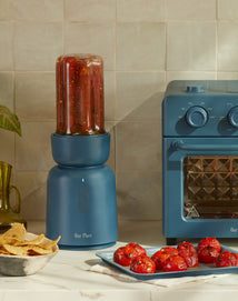 A blue blender filled with salsa stands next to a matching blue toaster oven; roasted tomatoes and a bowl of chips are on the counter.