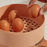 A hand places a brown egg into a peach-colored steamer tray with three other eggs already inside.