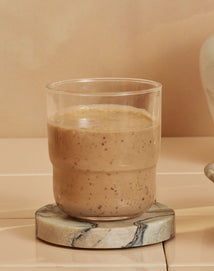 A clear glass filled with a thick, beige smoothie sits on a round stone coaster against a neutral background.