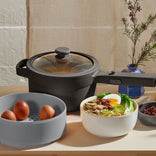 A black pot with a glass lid, a gray container holding four brown eggs, and a bowl of ramen with eggs and vegetables are arranged on a light wooden surface.