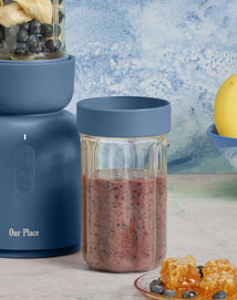 A blue blender with a glass jar of smoothie, a plate with honeycomb and blueberries, and a bowl with a banana are on a countertop.