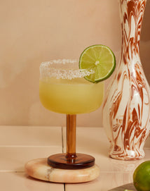 A margarita in a brown-stemmed glass with a salted rim and lime wheel garnish sits on a marble coaster beside a marbled vase.