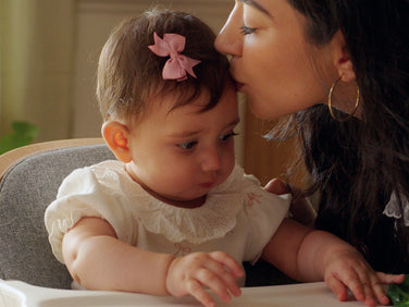 A woman kisses her baby on the forehead while the baby sits in a high chair.
