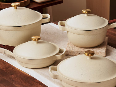 Four cream-colored pots and pans with gold knobs and wooden handles are arranged on a wooden kitchen counter with a runner and a hot pad.