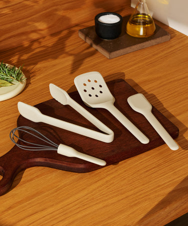 A whisk, tongs, slotted spatula, and mini spatula arranged on a wooden cutting board, with a small dish of herbs and peppercorns nearby.
