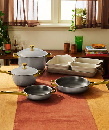 A set of gray cookware including a lidded Dutch oven, a lidded saucepan, two frying pans, and three rectangular baking dishes, all with gold-toned handles and knobs, arranged on a wooden table.