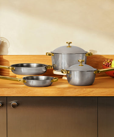 A set of grey cookware with gold handles and lids arranged on a wooden dining table.