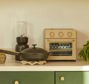 A beige toaster oven, a black lidded pan on a trivet, a blender, a striped cup, and a potted plant sit on a white kitchen counter with green drawers below.