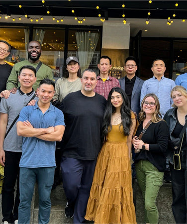 A group of sixteen people pose together outside a restaurant under string lights, smiling at the camera.