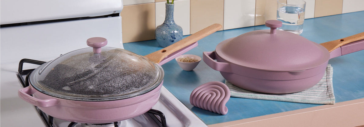 Translation missing: en.Two lavender-colored pans with wooden handles are on a blue kitchen counter next to a stove; one has a glass lid, and kitchen accessories are nearby
