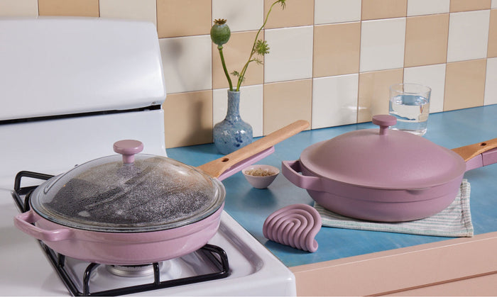 Translation missing: en.Two pink pans, one on a stove with a glass lid, the other on a blue countertop with a wooden handle, next to a glass of water, a vase, and kitchen accessories