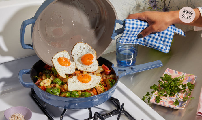 Translation missing: en.A person adds three fried eggs to a pan of cooked vegetables on a stove, using a blue checkered cloth to hold the pan lid