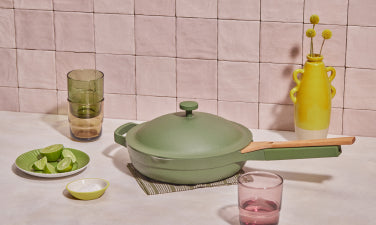 Translation missing: en.A green lidded skillet with a wooden handle sits on a placemat, surrounded by glasses, a yellow vase, and a plate of lime wedges on a tiled surface