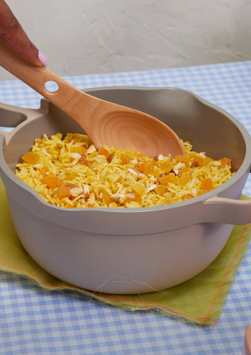 A hand stirs a pot of yellow rice mixed with chopped dried fruit and nuts using a wooden spoon on a checkered tablecloth.