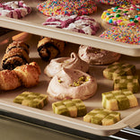 Two baking sheets filled with various cookies, including sprinkle-covered round cookies, pastries, meringues, and checkerboard shortbread squares.