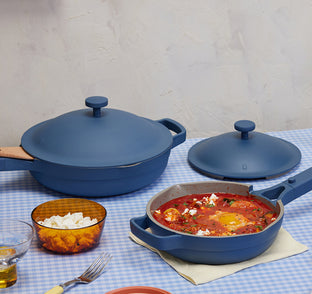 A blue nonstick pan with tomato stew on a checkered tablecloth, next to a matching pot with lid, a bowl of cheese, a glass of water, a fork, and a napkin.