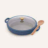 The Dual Handle Always Pan in blue, featuring a nonstick surface, stylish gold knob, and glass lid, is displayed beside a wooden spoon on a plain white background. This non-toxic cookware seamlessly blends style with functionality for effortless cooking.