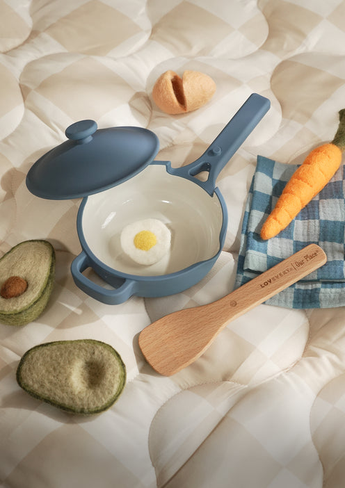 A toy blue saucepan with a lid, wooden spoon, and felt play food including an egg, avocado, and carrot on a checkered mat.