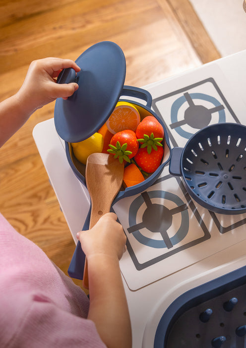 A child plays with a toy kitchen set, stirring plastic fruit in a blue pot with a wooden spatula.