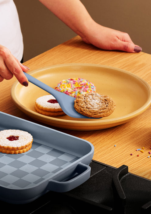A spatula lifts a cookie from a yellow plate, which holds other cookies, next to a blue baking pan.