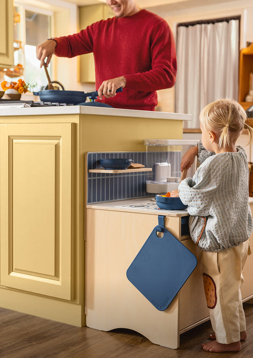 A man cooks at a kitchen counter while a young child plays at a nearby toy kitchen.