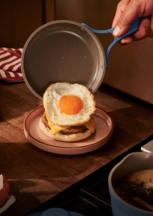 A hand places a fried egg from a pan onto a breakfast sandwich with cheese and sausage on a plate.