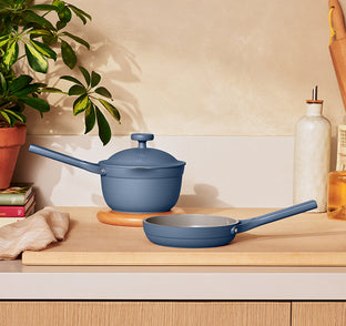 A blue saucepan with lid and a blue frying pan sit on a wooden kitchen counter next to a plant, books, a towel, and a bottle of oil.