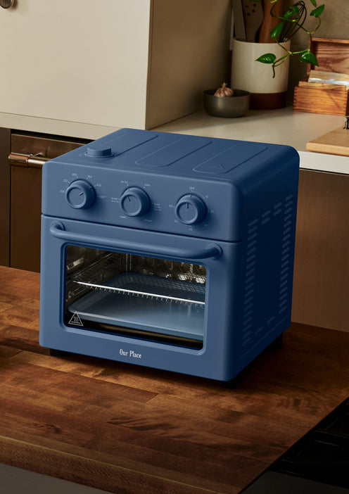 A blue countertop air fryer oven with three control dials sits on a wooden kitchen counter near a wall and some plants.