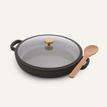 The Dual Handle Always Pan, featuring a black round design with a glass lid and gold knob, is paired with a wooden spoon and placed on a light background.