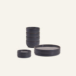 The Plates & Bowls Set features matte dark gray handcrafted ceramic plates, bowls, and four small dishes displayed on a light background.