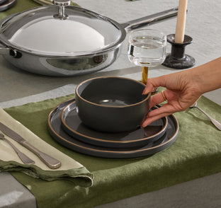 A hand places a dark ceramic bowl on a matching plate set beside a glass of water, cutlery, and a candle on a green and gray tablecloth.