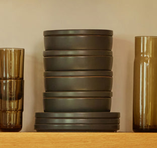 A stack of dark ceramic bowls and plates is placed between two tall amber-colored glass cups on a wooden shelf.