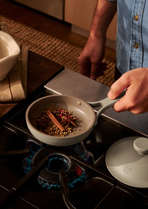 A person holds a small pan with spices, including cinnamon sticks and star anise, over a lit gas stove burner in a kitchen.