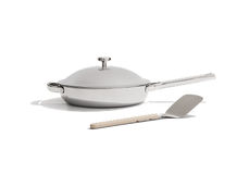 A stainless steel frying pan with a lid and a metal spatula with a light-colored handle, set against a plain white background.