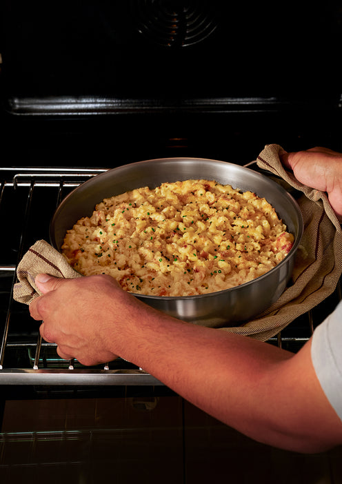 Person placing a round pan of baked macaroni and cheese with a golden crust into an oven, using a towel to hold the hot pan.